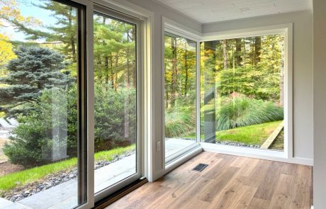 Window remodeling project featuring large floor-to-ceiling sliding glass windows with white trim, hardwood floors, and panoramic views of a landscaped wooded backyard by Craftworks Construction.