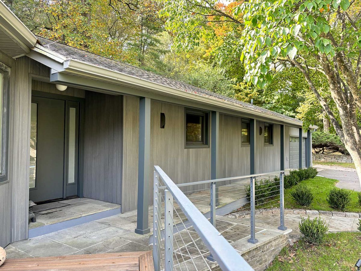 Home remodeling project featuring a modern ranch-style exterior with gray vertical wood siding, cable railing, stone patio, and updated windows surrounded by mature fall foliage.