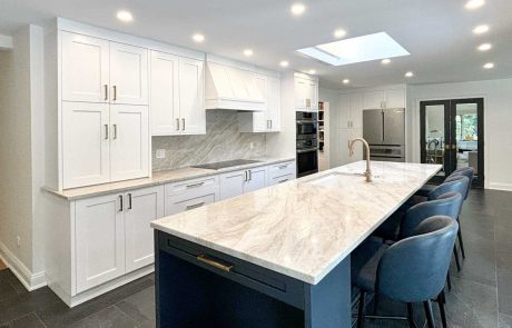 Kitchen remodeling project featuring white shaker cabinets, navy blue island with marble countertop, gold hardware, blue leather bar stools, skylight, and dark slate tile floors.
