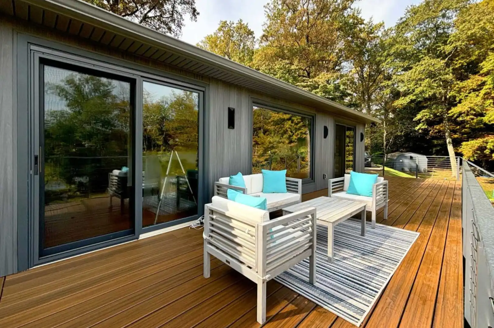 Deck remodeling project featuring a wide hardwood deck with outdoor lounge furniture, teal accent pillows, large sliding glass doors, gray wood siding, and waterfront wooded views by Craftworks Construction.