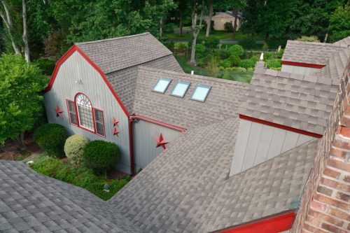 Roofing remodeling project in Fairfield, CT featuring newly installed brown architectural shingles, skylights, and red trim accents on a gray barn-style home surrounded by mature landscaping.