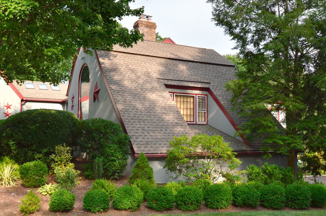 117 Roof remodeling project on a gray cottage-style home with newly installed gray shingles, steep roofline, red trim accents, and brick chimney surrounded by mature landscaping.