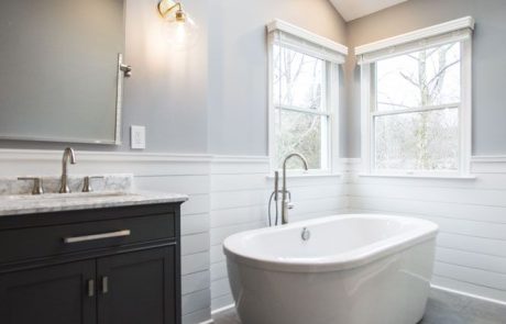 Renovated bathroom with freestanding soaking tub, shiplap walls, dark vanity with marble countertop, gray tile floor, and skylight.