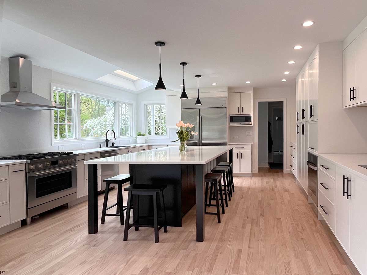 Modern renovated kitchen with white shaker cabinets, large dark island with black bar stools, stainless steel appliances, pendant lights, and light hardwood floors.