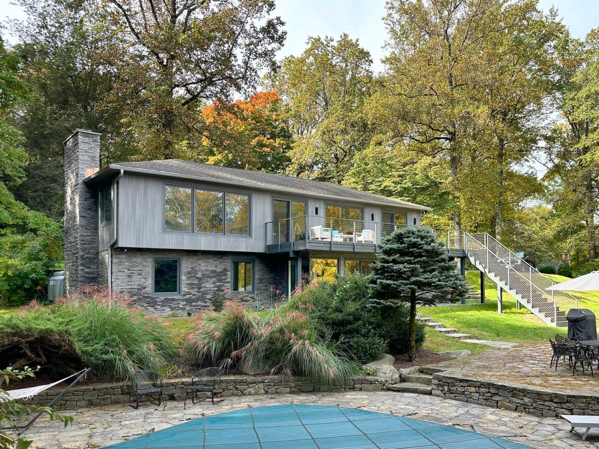 Home remodeling by Craftworks Construction LLC featuring a modern split-level home with gray wood siding, stone facade, large windows, cable railing balcony, and pool patio surrounded by fall foliage.