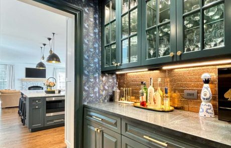 Home remodeling project featuring a dark green butler's pantry with glass-front cabinets, copper tile backsplash, brass hardware, and hardwood floors opening to a renovated kitchen.