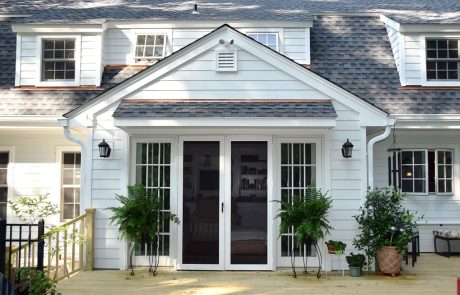 Home remodeling project featuring new white vinyl siding, French doors, updated windows, gray shingle roof, and classic wall lanterns on a colonial-style home exterior.
